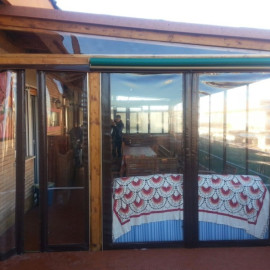 Veranda in legno con tamponature in PVC Cristal trasparente. Realizzato in un attico a Ostia (Roma). Chiusure laterali apribili con stecche di rinforzo, progettata per resistere al vento.